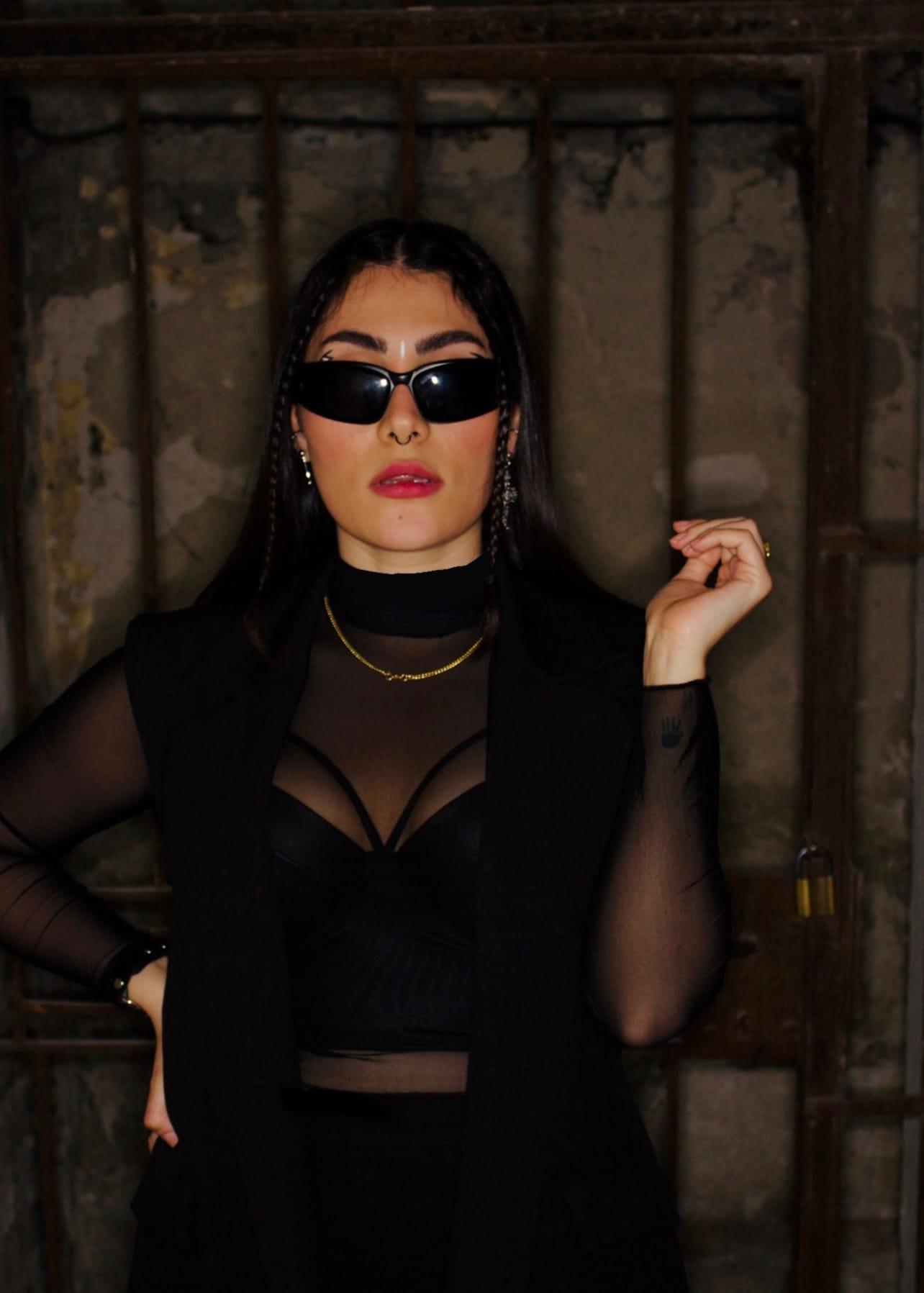 5OGOL in sunglasses and mesh top against a rusted wall
